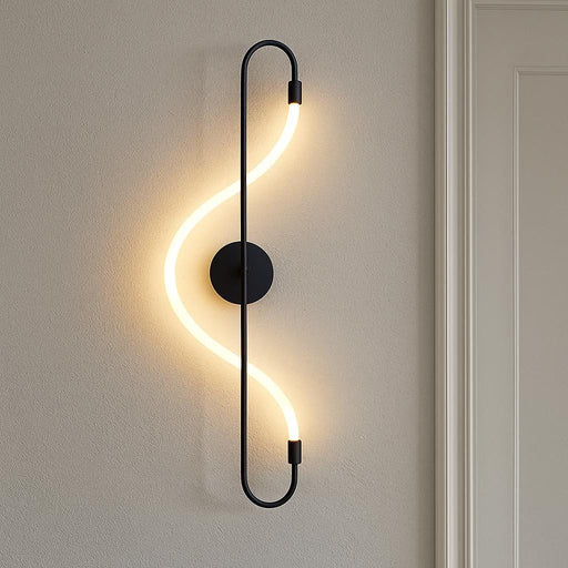 A studio shot of the Iris Wall Light in matte black, installed in a contemporary hallway with minimalist decor, showcasing its fluid S-shaped form and the warm white light illuminating the textured wall behind it.