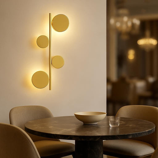 A modern dining area featuring the Sam Gold Wall Light installed above a stylish dining table, showcasing the sculptural design and the warm white glow illuminating the space, with a blurred elegant background.